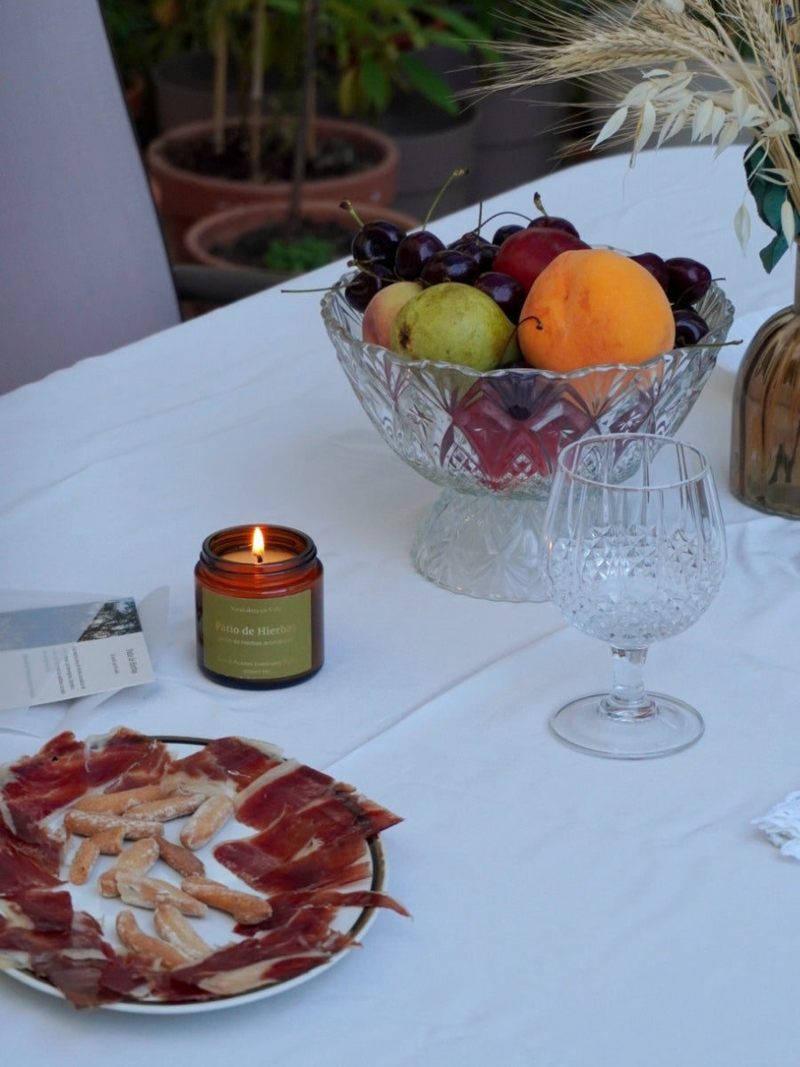 Vela Patio de Hierbas de Naturaleza en Vela en mesa de jardín con mantel blanco, flores y copa de cristal al fondo